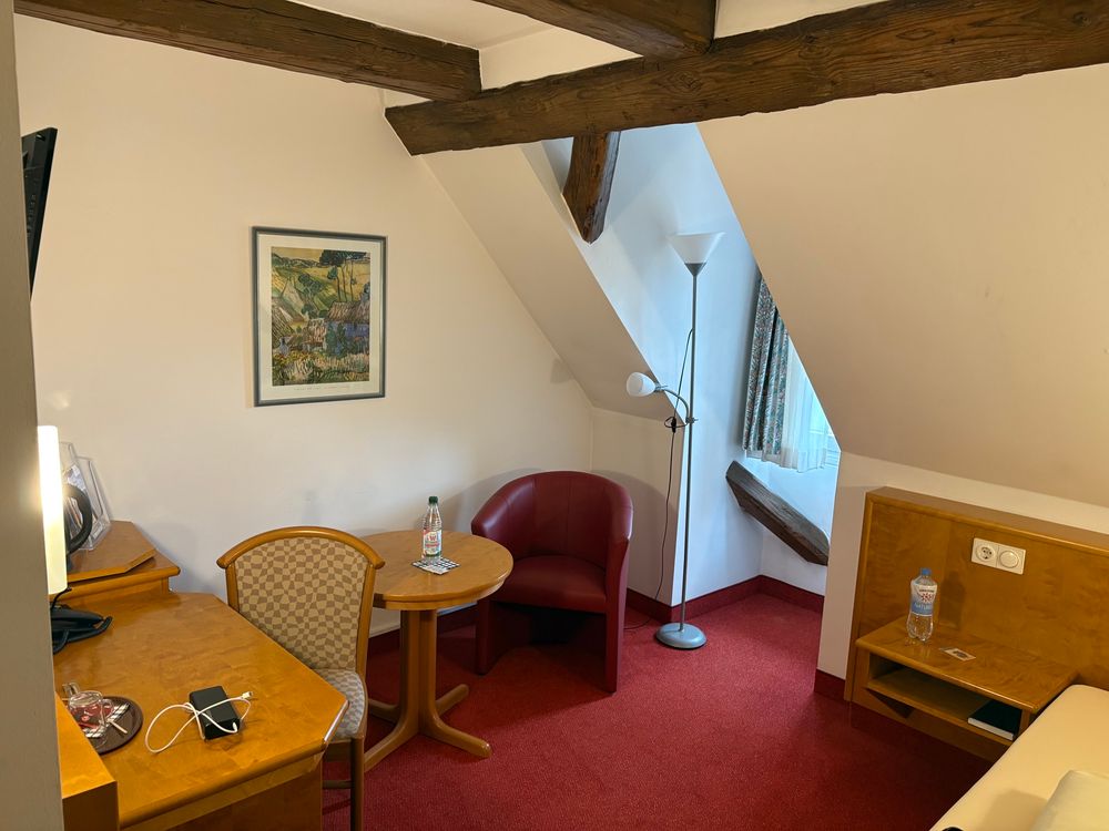 Blick in ein Dachboden-Hotelzimmer mit Fachwerk an der Decke. Es wirkt klein, mit einem Bett rechts, links ein Schreibtisch mit Stuhl, zentral hinten an der Wand ein kleiner runder Tisch und ein Sessel mit halbhoher Lehne. Durch ein winziges Dachfenster fällt etwas Licht herein. 