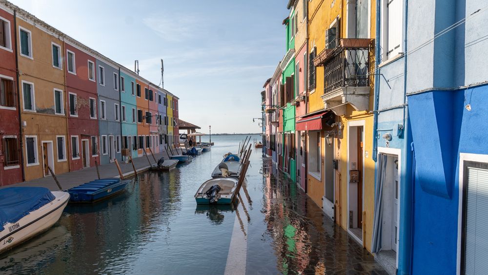 A sunny canal on the island of Burano (Venice), lined with two rows of vibrantly colored houses (blue, yellow, red, green). Small boats are moored in the canal, and the water is high enough to slightly flood the walkways in front of the doors (Acqua Alta).