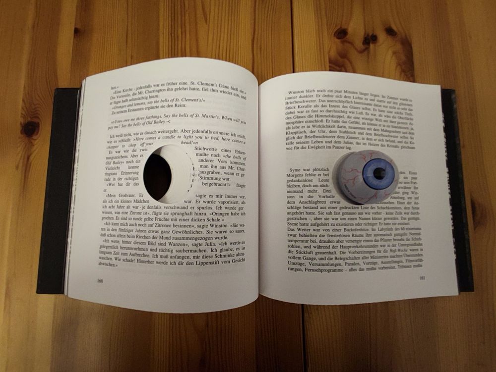 Das Buch liegt aufgeschlagen auf einer Holzoberfläche. Auf der rechten Seite des aufgeschlagenen Buches ist wieder das künstliche Auge zu sehen, das nun auf dem bedruckten Text der Buchseite liegt. Auf der linken Seite der linken Buchseite ist ein großes, kreisrundes Loch zu sehen, das durch die Seiten führt. Der Text im Buch ist in deutscher Sprache.