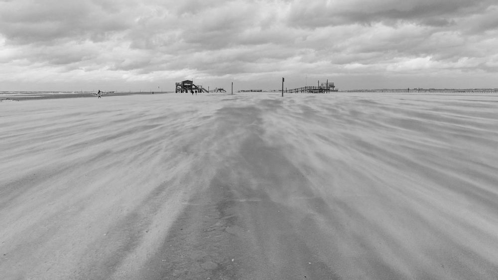 Ein Schwarz-Weiß-Foto zeigt einen breiten Sandstrand, auf dem der Wind Sandwirbel über die Fläche treibt. Im Hintergrund sind auf Holzpfählen mehrere Gebäude zu sehen, die in der Ferne verschwinden. Der Himmel ist bedeckt mit dichten Wolken.
