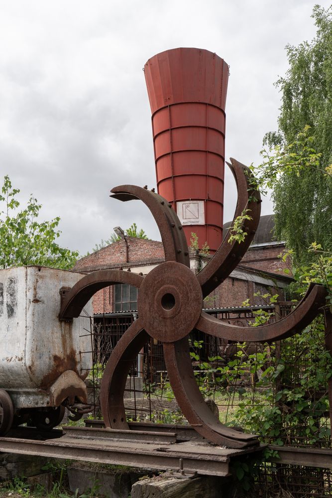 Ein großes, verrostetes Schaufelrad aus Metall steht im Vordergrund. Dahinter befindet sich ein weißer, kastenförmiger Wagen auf Schienen. Im Hintergrund ist ein hohes, rotes, konisch zulaufendes Metallrohr zu sehen, das an einem Backsteingebäude befestigt ist. Die Szenerie ist von Pflanzen überwuchert und der Himmel ist bedeckt.
