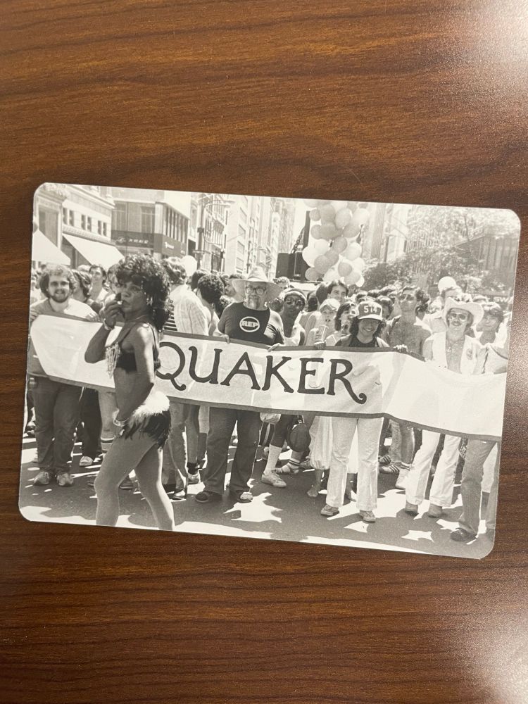 Black and white photograph of people carrying a banner that says QUAKER, labeled on back “Friends with banner in gay pride parade, up 5th avenue, early 1980s”
