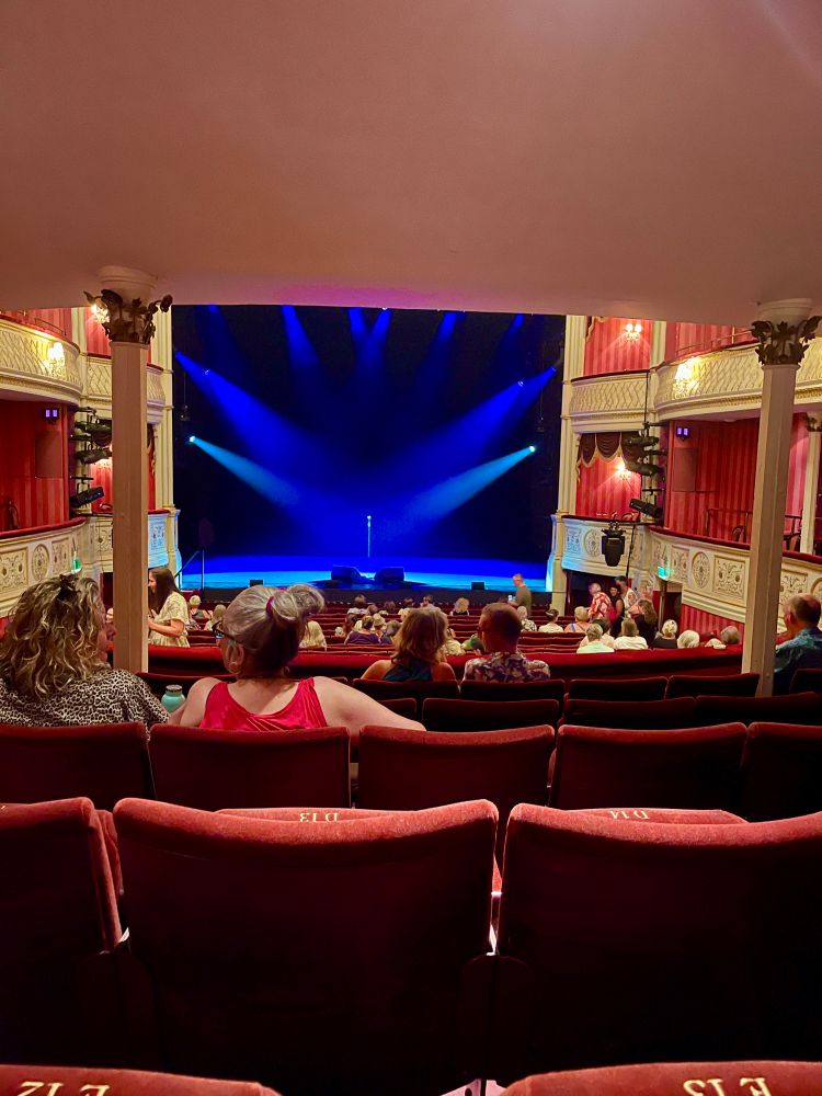 View of the stage at the theatre royal Bath from the circle. Dark blue light beams cross the stage highlighted by haze. Downstage light blue beams highlight the microphone.