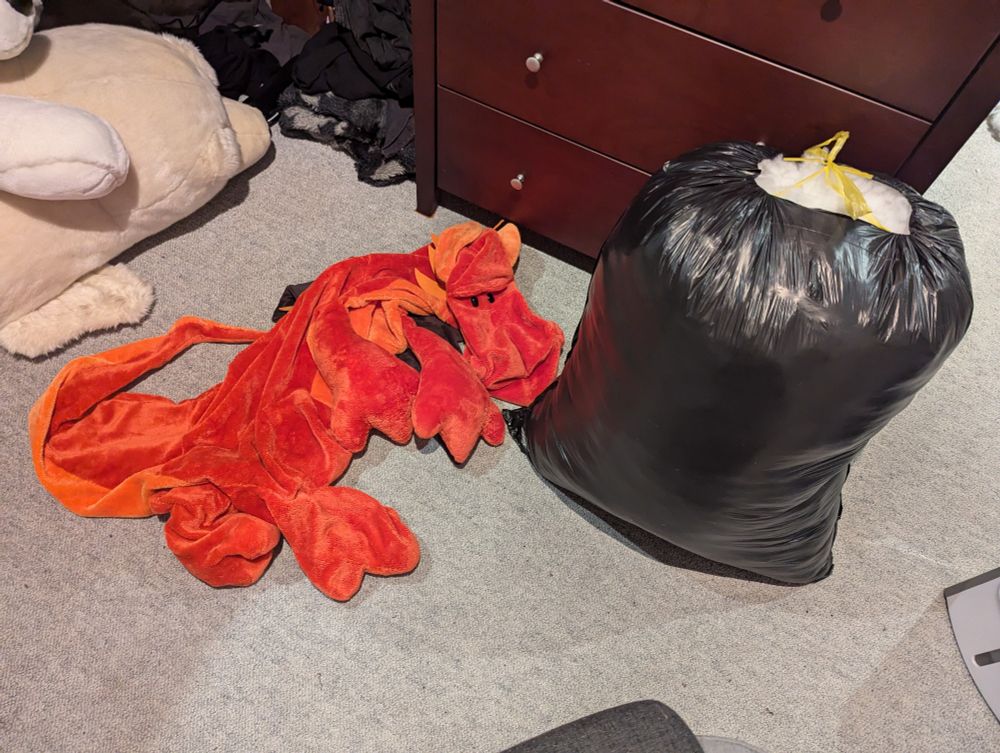 Large Unstuffed dragon plush laying on the floor next to a garbage bag full of stuffing. 