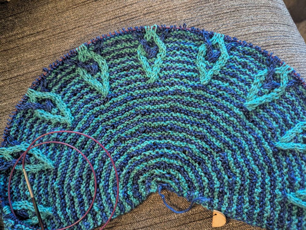 Slightly more than half pi shawl in blue and seafoam green stripes with the beginnings of 9 interlinked cables around the upper outside curve.