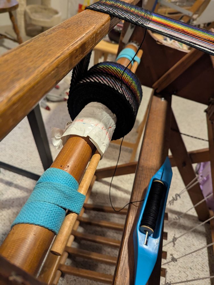 The front beam of a floor loom, holding a warp that is 2x2 twill, in the colors of the Pride Progress flag. Black weft yarn is on a blue boat shuttle.