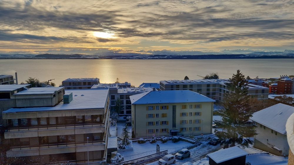 View of the Neuchâtel lake in Switzerland. The sun is shining through a thin cover of clouds and reflects on the lake. Alps in the distance. Buildings in the foreground and landscape in the background are all covered by a thick layer of snow. 