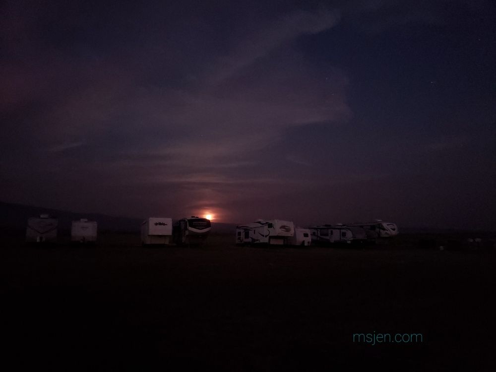 Second photo of the smokey skies of August: One evening past full Moon rising over the trailer storage area. The sky is dusky red and purple with clouds and wildfire smoke. The partially risen Moon is large on the horizon and dark red. The foreground is dark with a few campground trailers in the fading light. Photo by Jenifer Hanen, taken on Aug 9, 2025.
