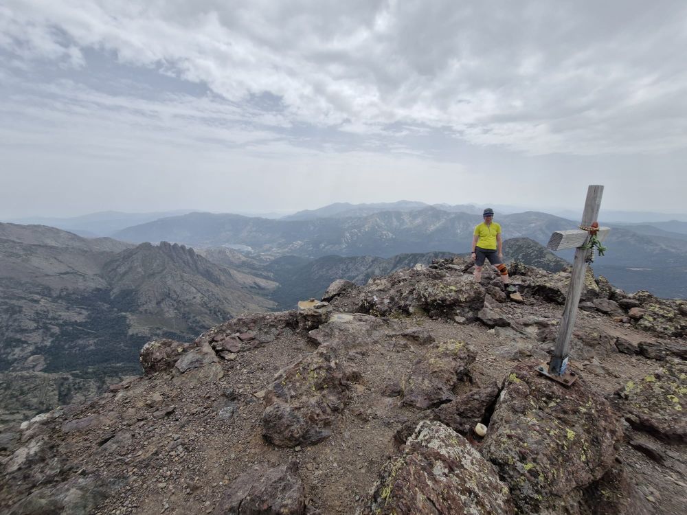 Women in yellow top with dodgy knee on top of high mountain with a cross in the foreground and surrounding hill beyond.