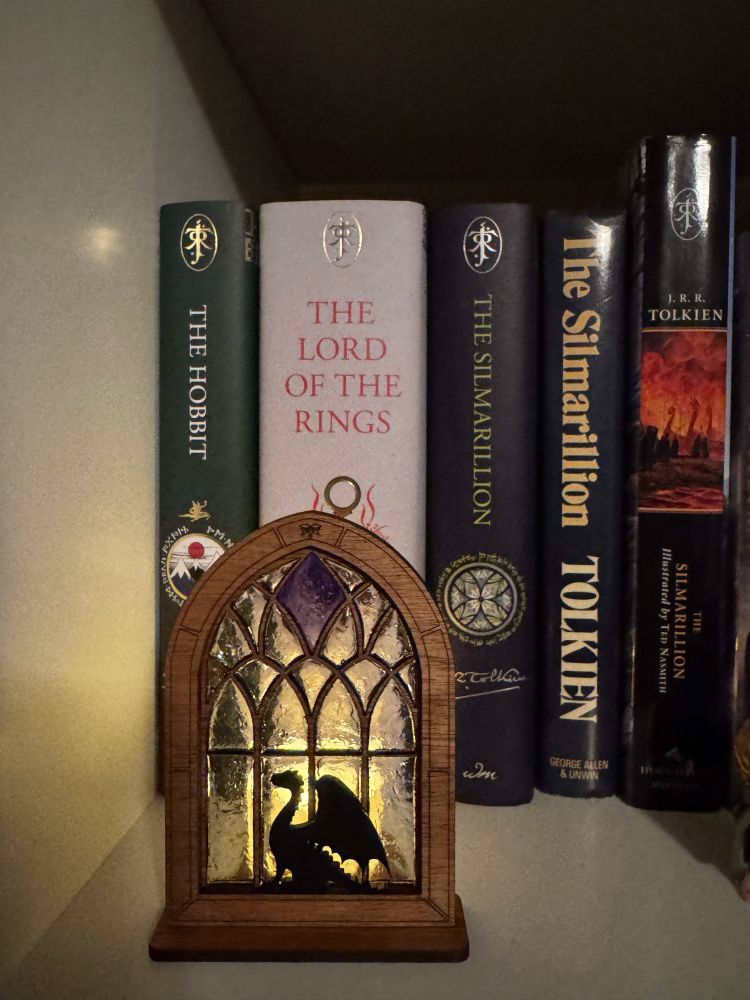 A close up of a white bookshelf with decorative window with a dragon sitting in front of it, a fake candle behind it casting a warm glow through the window panes. In the background are copies of The Hobbit, The Lord of the Rings, and three copies of The Silmarillion in different editions.