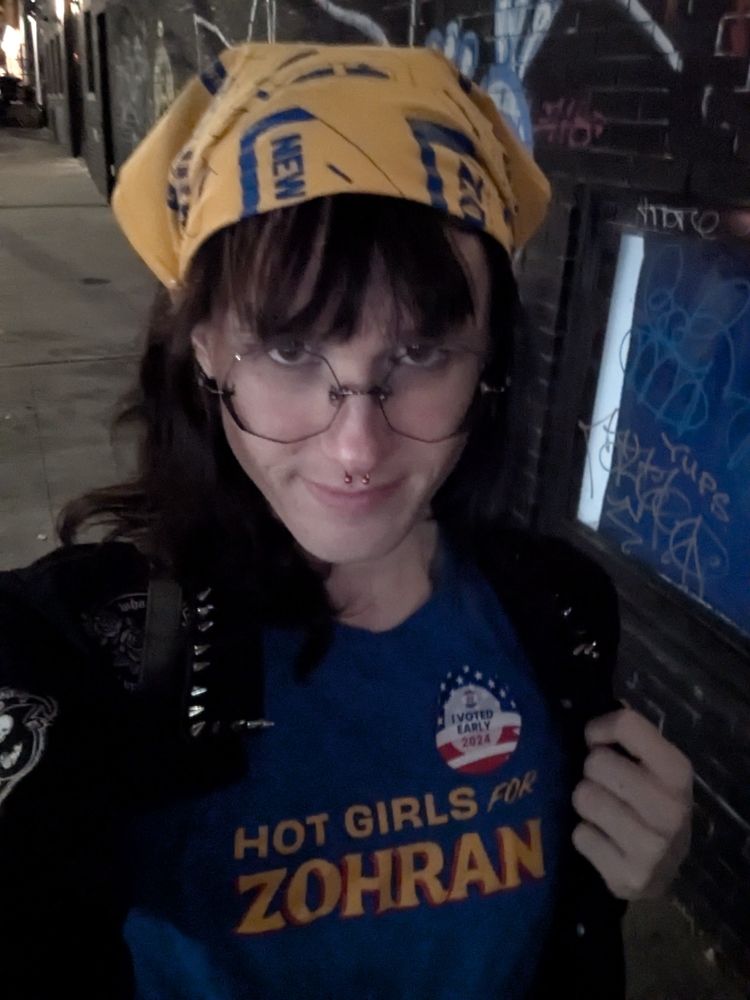 me walking down a street wearing a Hot Girls for Zohran short and bandana with an I Voted sticker 