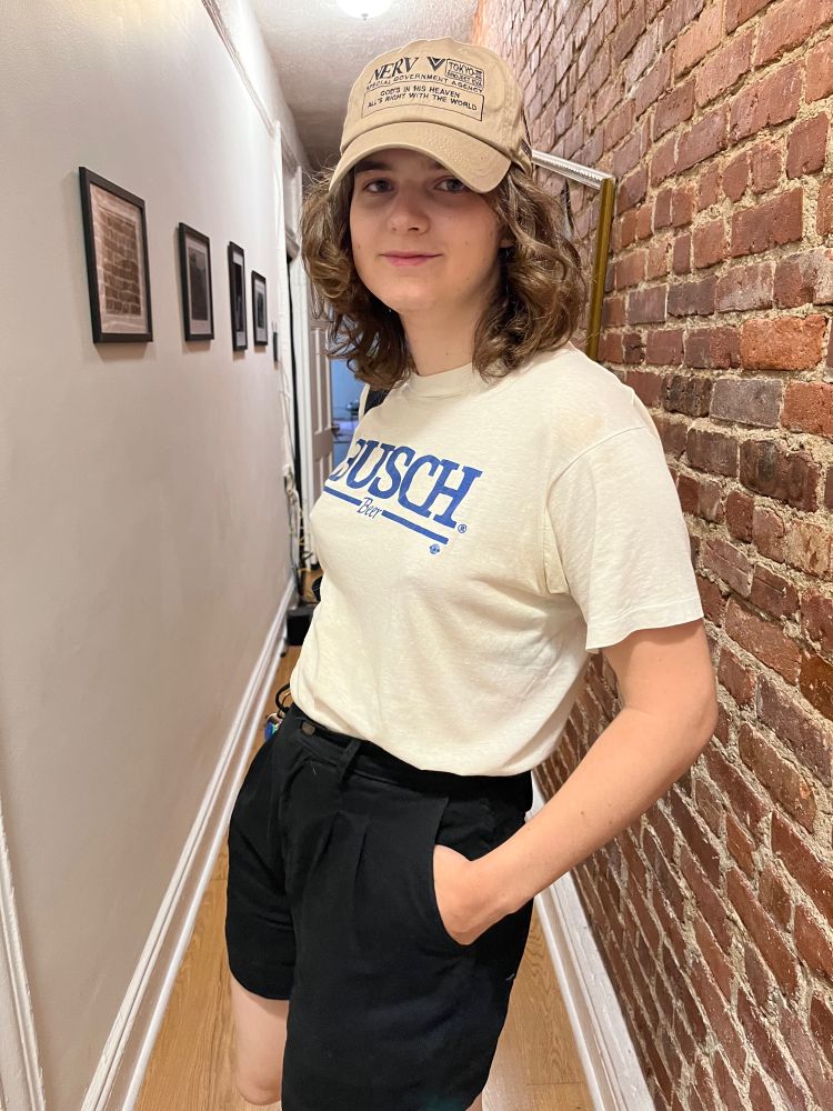 Sloane in hallway of apartment wearing Busch Beer t shirt and Neon Genesis Evangelion hat. She looks really cute.