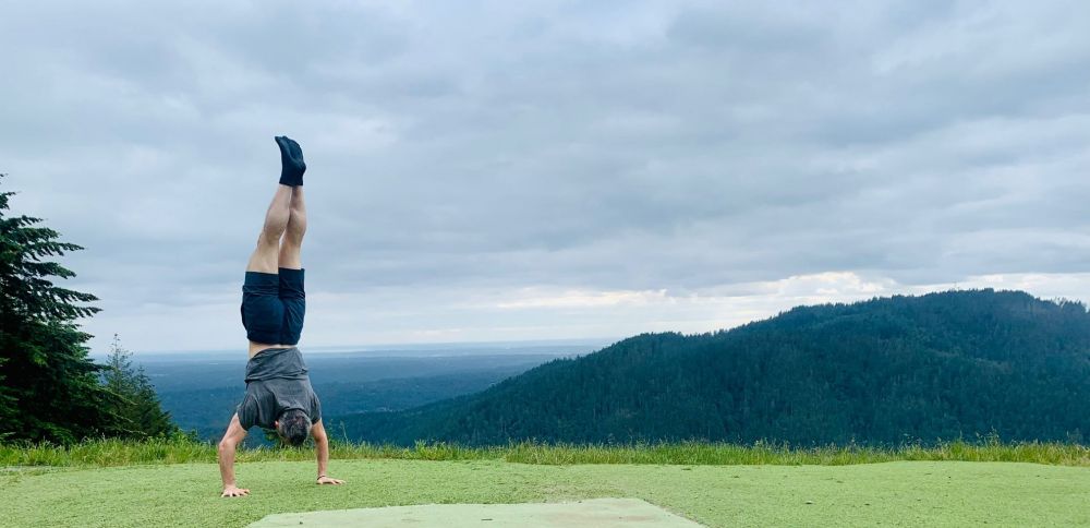 Yours truly doing a handstand in a gray shirt and blue shorts on a patch of grass. Fores-covered mountain in the background