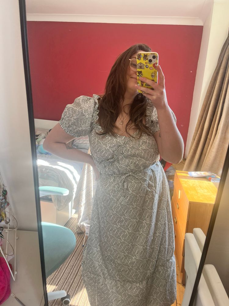 I’m taking a mirror selfie wearing an across body dress toed with a ribbon I made. The pattern is small lavender sprouts on a cream background repeated a lot! The phone I’m using is covering half my face.