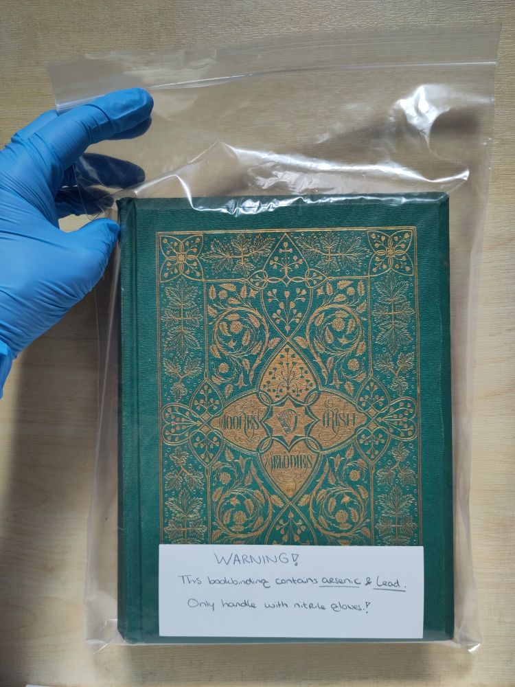 Photograph of an hand inside a blue nitrile glove holding the book entitled Moore's Irish Melodies which is inserted into an archival-quality zip lock bag. A handwritten note on the front reads: 'WARNING! This bookbinding contains arsenic & lead. Only handle with nitrile gloves.'