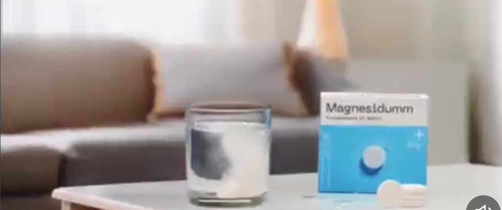 Wasserglas gefüllt auf Tisch, daneben blau/ weiß gestreifte Tablettenpackung mit der Aufschrift „Magnesidumm“. Im ahintergrund ein graues Sofa.