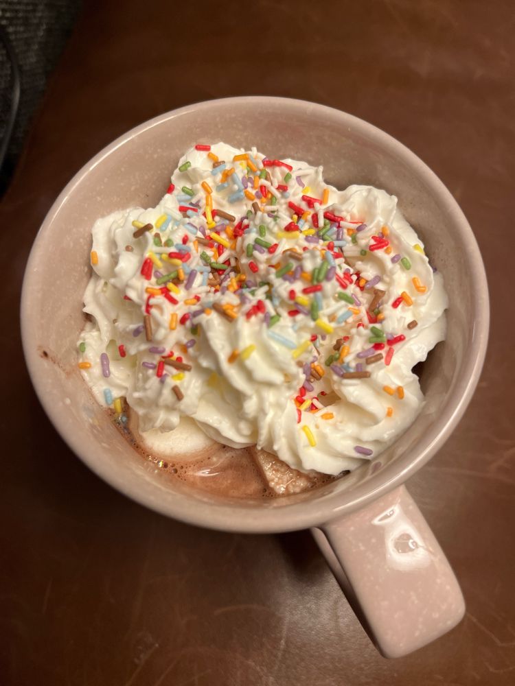 Sofalehne (braun) mit rosa Tasse.
Bunte Zuckerstreusel auf Schlagsahne - darunter Marshmallow und Kakao.☕️