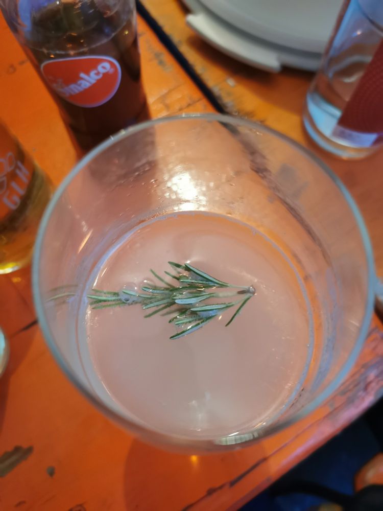 Weinglas von oben mit alkoholfreien Rhababer Spritz und einem Rosmarinzweig. Es steht auf einer orangen Bierbank