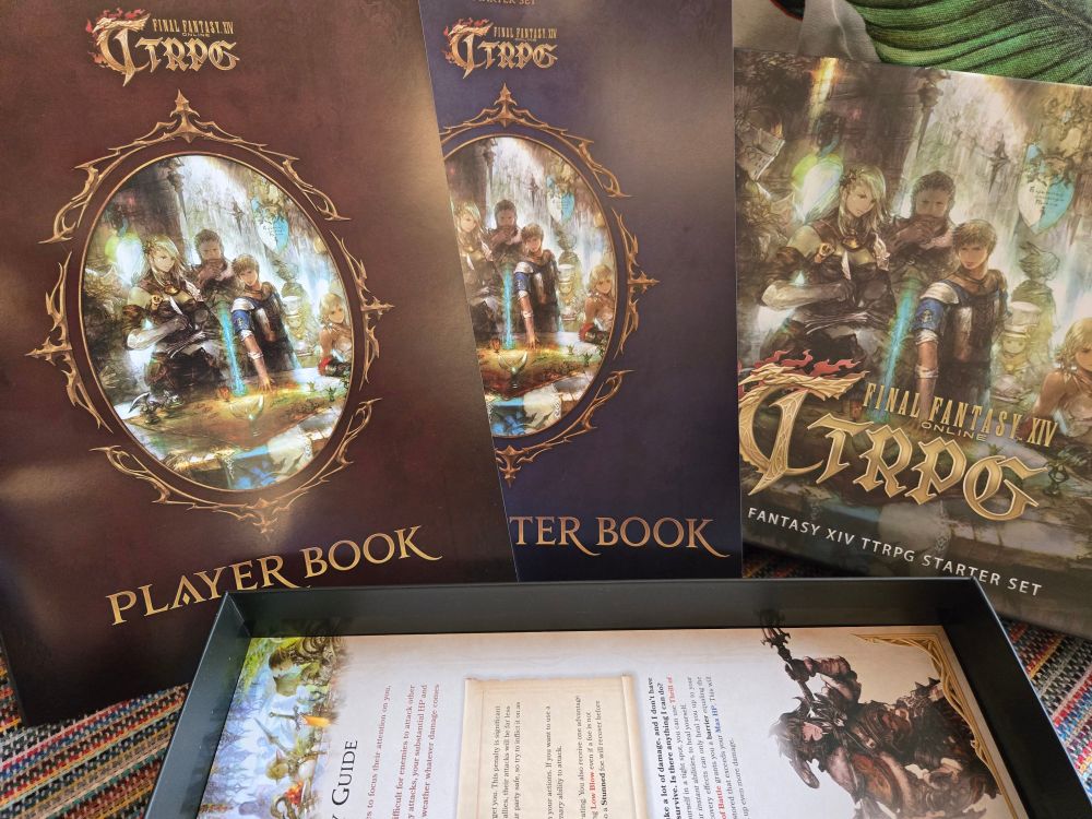 A imagem mostra o conteúdo do Final Fantasy XIV Online TTRPG Starter Set disposto sobre uma superfície. No centro, estão dois livros com o título "Player Book" em letras douradas na parte inferior, com capas que apresentam uma arte detalhada de personagens de Final Fantasy XIV dentro de uma moldura decorativa dourada. À direita, a caixa do Starter Set está visível com uma arte semelhante à capa principal do conjunto, destacando um grupo de aventureiros. Na parte inferior, dentro da caixa, é possível ver parcialmente um guia de introdução com ilustrações vibrantes de personagens e ambientes do universo de Final Fantasy XIV. A composição destaca o conteúdo premium do conjunto e evoca o espírito do RPG de mesa.

