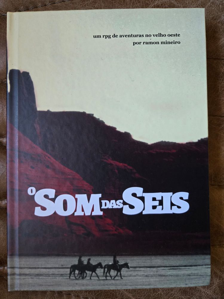 A imagem é a capa do livro "O Som das Seis", um RPG de aventuras no Velho Oeste criado por Ramon Mineiro. A capa apresenta uma fotografia de uma paisagem típica do oeste americano, com formações rochosas ao fundo e silhuetas de cavaleiros montados em seus cavalos em primeiro plano, criando uma atmosfera de exploração e aventura característica do gênero faroeste.
