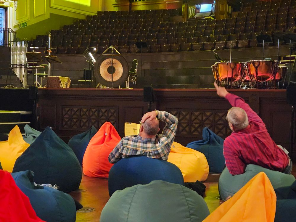 People sitting on brightly colored beanbag chairs in front of a stage with tubular bells, a gong and kettle drums but no performers yet.