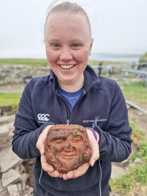 Student Katie Joss standing in front of an archeological site in Orkney, grinning and holding in her open palms a red sandstone head.   From here:

https://www.smithsonianmag.com/smart-news/archaeology-student-discovers-carefully-carved-stone-head-at-viking-settlement-in-scotlands-orkney-islands-180987111/