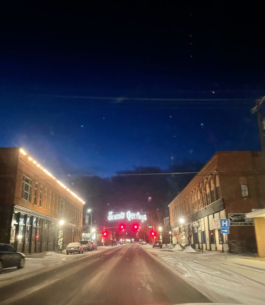 Holiday lights over an intersection in a wintry small Montana town that say “seasons greetings” 