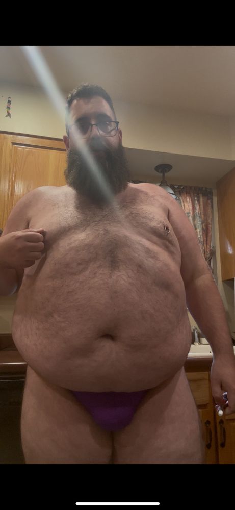 A bear tweaking his nipple in a purple thong and a cloud of smoke.