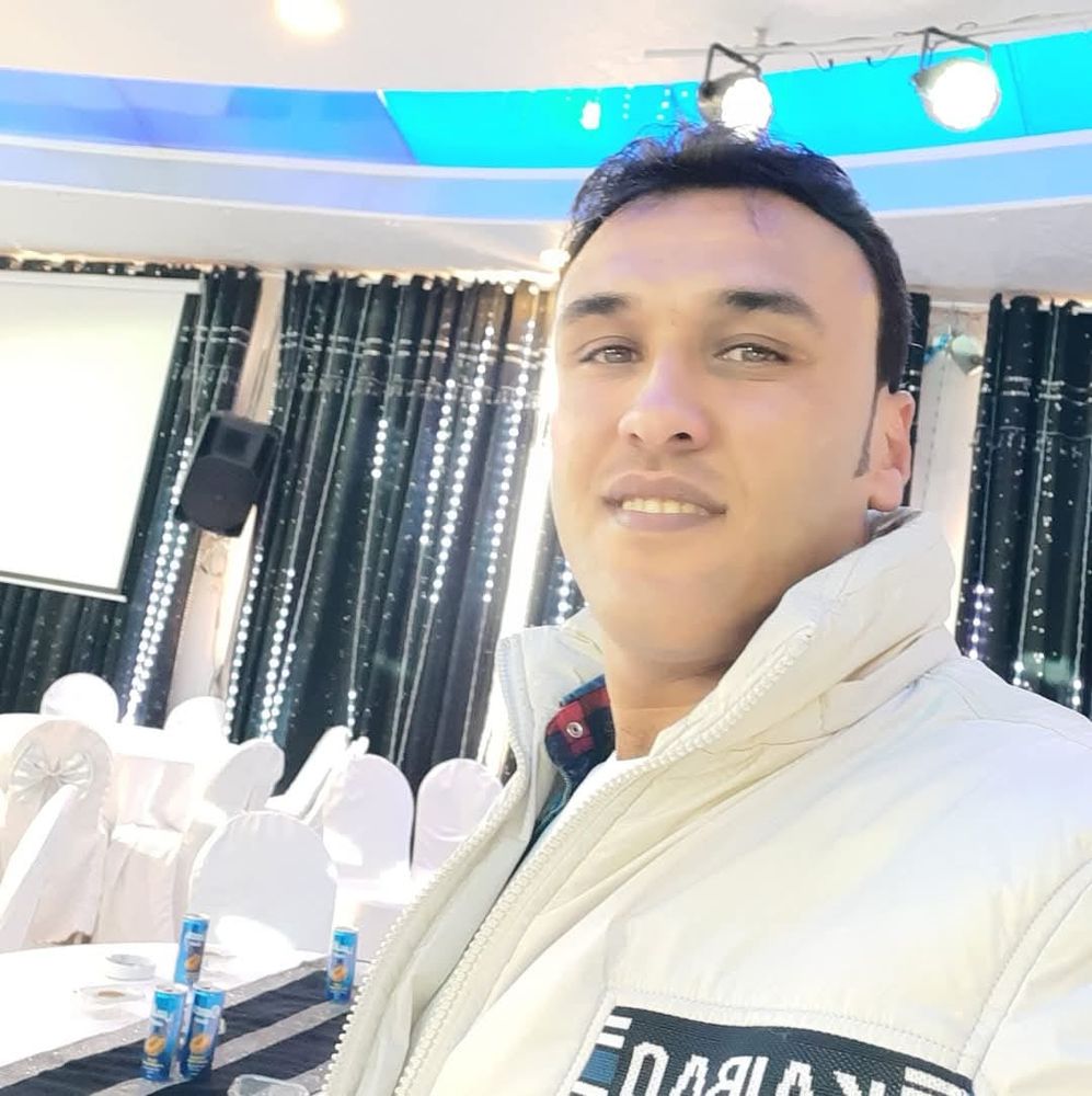 a man wearing a white jacket, Magdy, smiles at the camera, in a room with curtains in the background