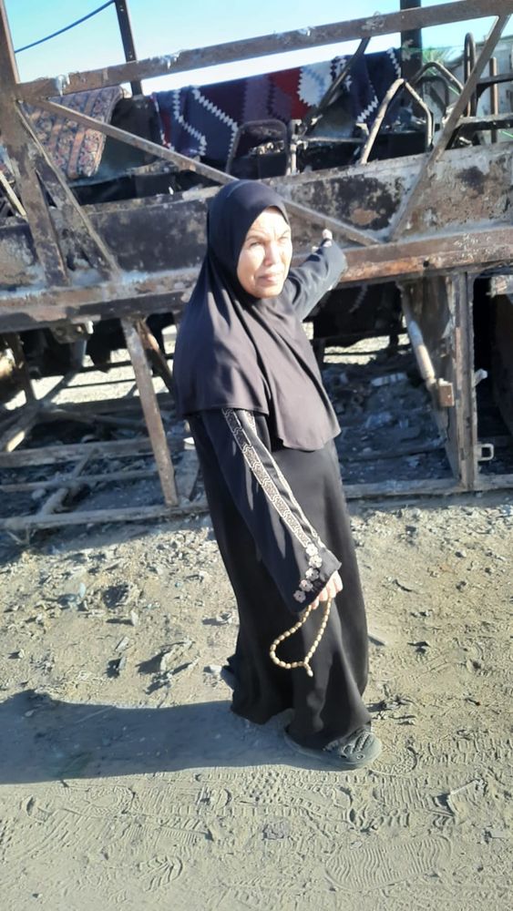 a woman, Fatima, stands outdoors wearing a black dress and headscarf, gesturing to the metal skeleton of a bus destroyed by a bomb behind her