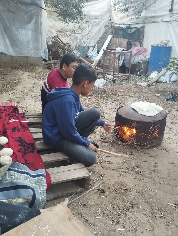 two boys, Moaz and Karam, sit on a wooden palette and add sticks to a fire they are using to bake flat bread. Tents and remnants of furniture can be seen in the background