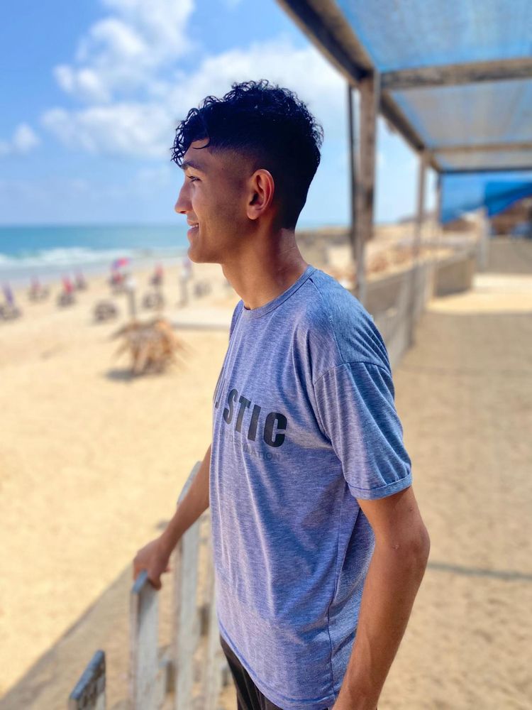 a teenage boy, Ahmed, is at the beach, wearing a t-shirt, smiling while looking toward the sea