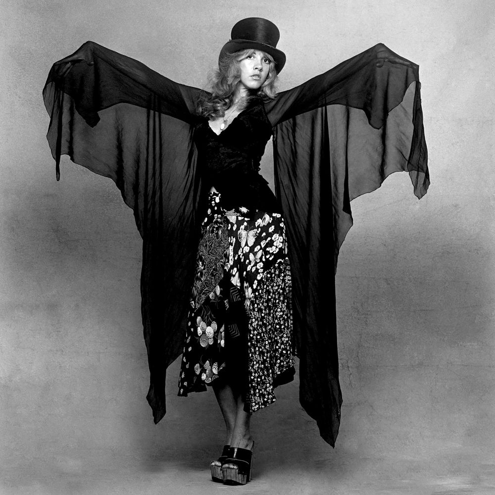 Black and white image of musician Stevie Nicks wearing a top hat and draped in a large black scarf that looks like butterfly wings
