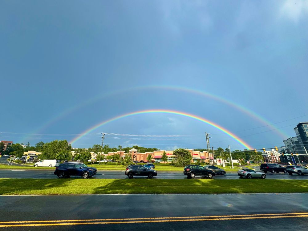 A double rainbow stretching above a typical American shopping center with a line of traffic in front of it. The inner rainbow is vividly intense, with the lines of color sharply defined, especially on the right side. The outer rainbow is less vivid, but still clearly visible. The space between the two rainbows is a dark purply color, giving the rainbow something of the feeling of a solid object. The shopping center behind the camera was destroyed by several feet of floodwaters a few days ago, and heavy rains have continued since. Meteorological phenomena do not actually symbolize anything, but it sure would be nice if this meant that the worst is over.