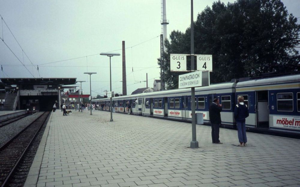 Das historische Bild zeigt den Olympia-Bahnhof mit einer alten S-Bahn mit blau-weißer Lackierung. 