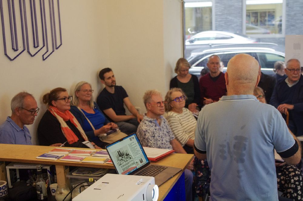 Man sieht die Rückseite eines Mannes, der einen Vortrag hält. Im Hintergrund sitzende Personen im Publikum in einem Ladenlokal. 