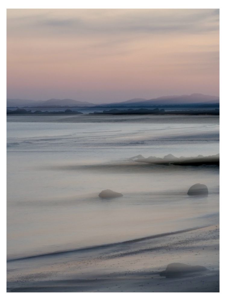 A pink sky as the sun sets over the sea. There are hills in the background and some rocks in the foreground on the beach.