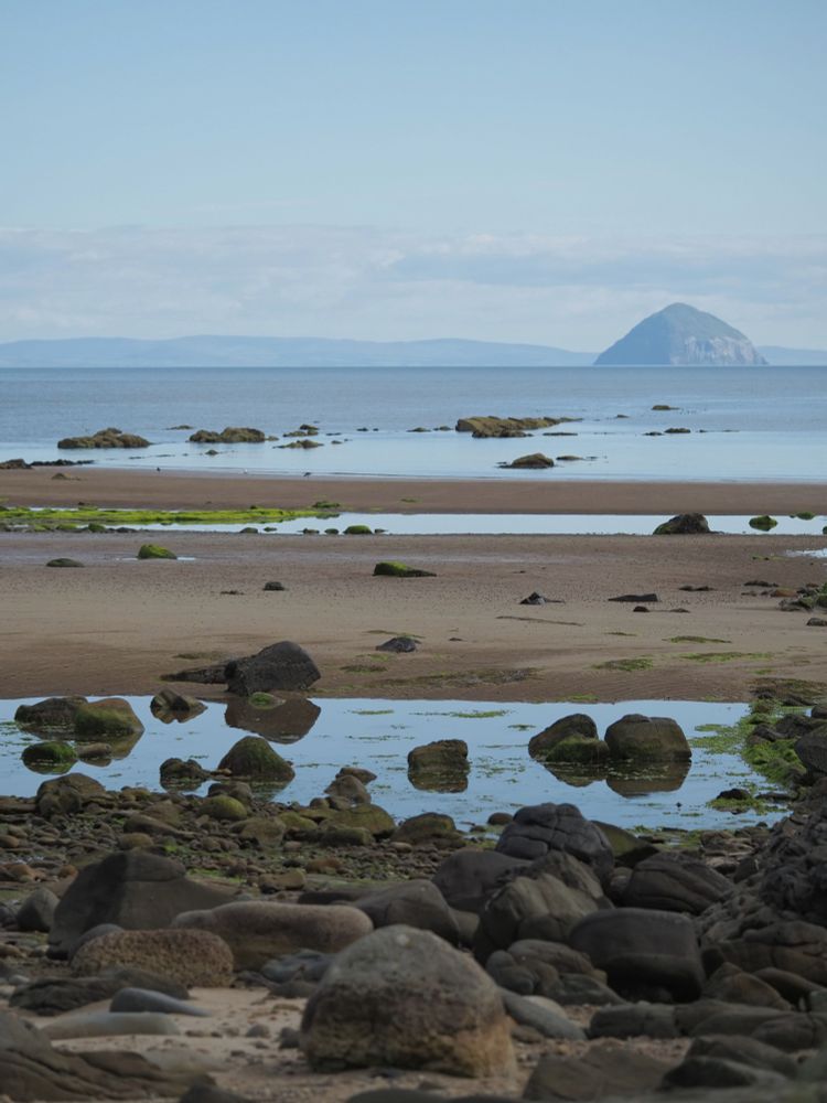 A view across rocks and a sandy beach to a  island in the sea. The sea and sky are pale blue, with a low band of white grey clouds in the sky.