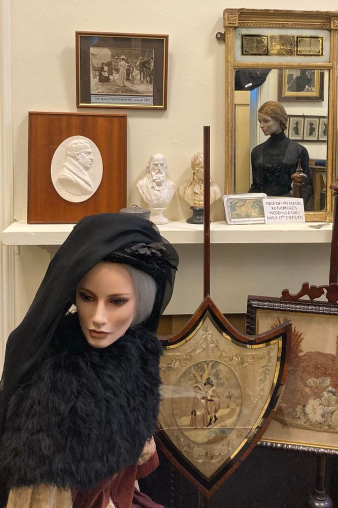 A tableau of museum objects, including two mannequins dressed in mourning wear (one glimpsed in a mirror), as well as photographs, silhouettes, busts, fire screens and a piece of Mrs Samuel Rutherford’s wedding dress (early 17th century).