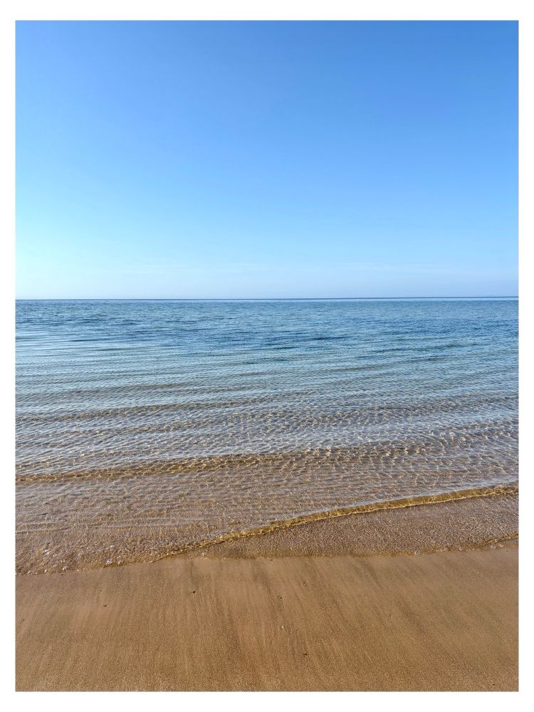 Blue sky over a blue sea with small clear ripples of water coming up onto golden sand.