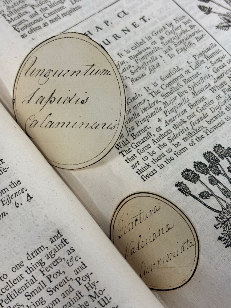 Two circular handwritten labels tucked inside a book, with the captions ‘Unguentum Lapidis Calaminaris’ and ‘Tinctura Valeriana Ammoniata’.