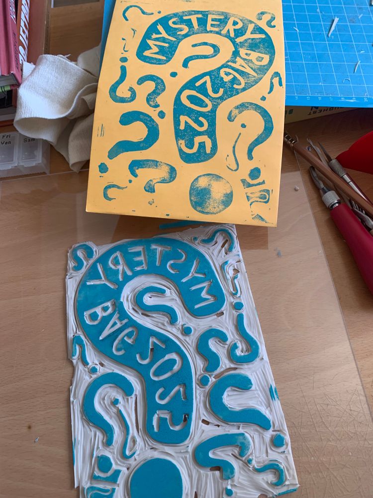 A carved Lino piece covered in question marks, the largest labeled “Mystery Bag 2025”