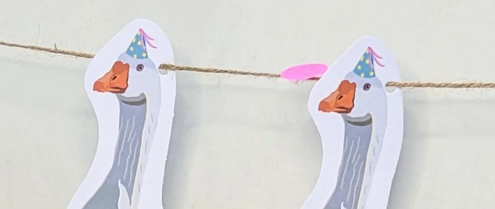 Card goose bunting, two goose heads and necks are visible, both are wearing party hats, rough string is strung through holes behind their heads 