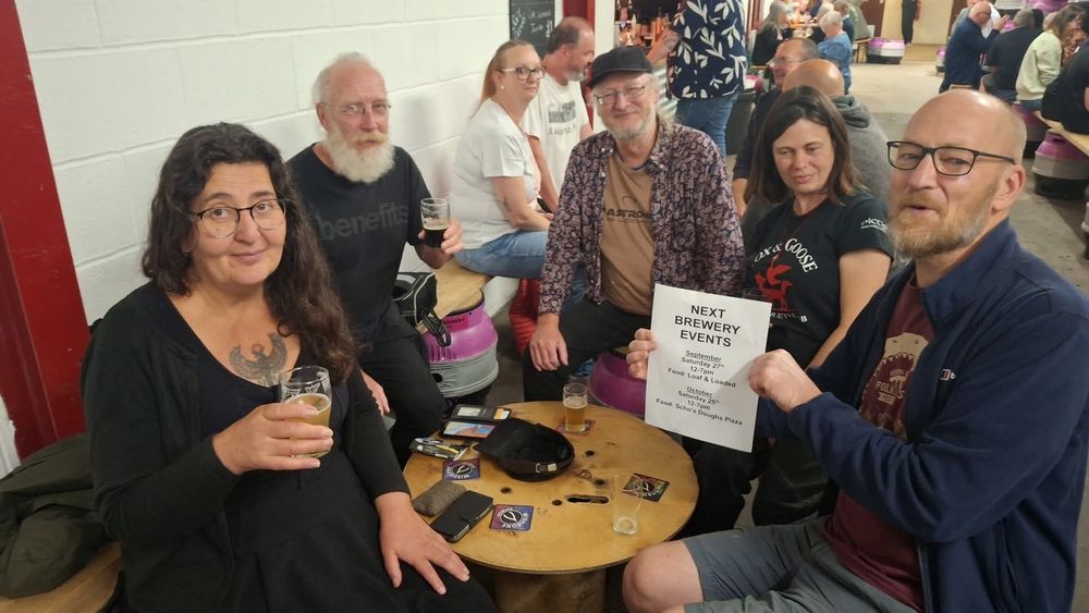 Five people sit around a small circular table, they are all drinking beer and seem pleased with themselves. One is holding up a piece of paper with the dates of the next brewery tap events.