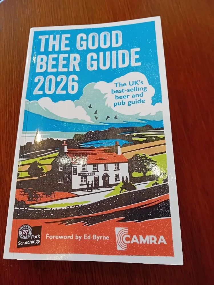2026's Good Beer Guide on a brown wooden table. This cover features a stylised country pub among green fields and under a blue sky. Looks pretty idyllic.