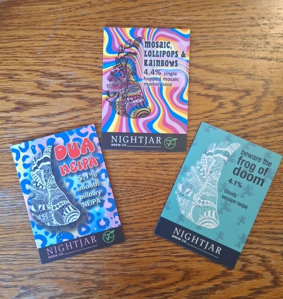 Three colourful Nightjar pump badges on a brown wooden table. Left, Dua Neipa, 5.1%, pink and blue leopard print background; centre Mosaic, Lollipops and Rainbows, 4.4, pink, blue, orange and yellow swirled background; and left, Beware the Frog of Doom, 4.1%, session NEIPA, light blue background. 