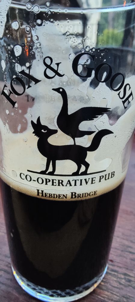 A half full/empty pint glass on a table. Its black beer is the same colour as the black logo of a stylised fox with a goose standing on its back. The logo text reads Fox & Goose, cooperative pub, Hebden Bridge.