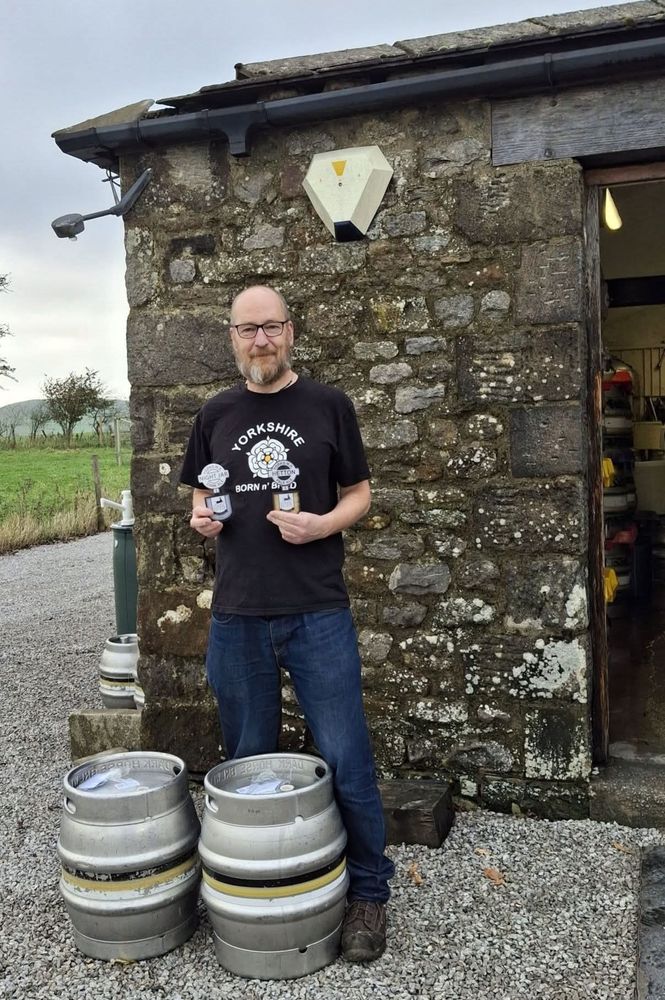 A balding middle-aged man in a black Yorkshire t-shirt and jeans stands outside a stone built building. At his feet, two beer kegs, he seems pleased with himself. 