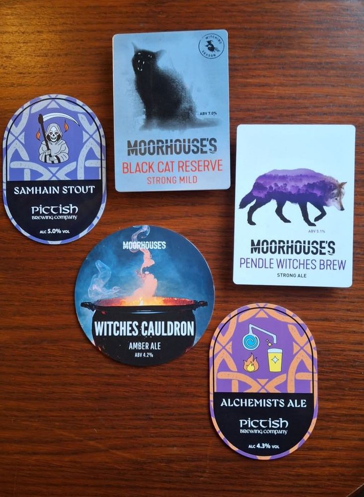 Five pump badges on a wooden table top. Pictish Samhain stout and alchemist's ale, and moorhouses black cat reserve, witches cauldron and witches brew. Featuring a selection of Halloween appropriate imagery. 