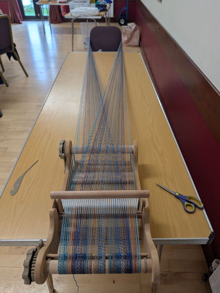 A rigid heddle loom warped up in muted blues, oranges, greens, stretched across a table with a pair of scissors and a threat. 